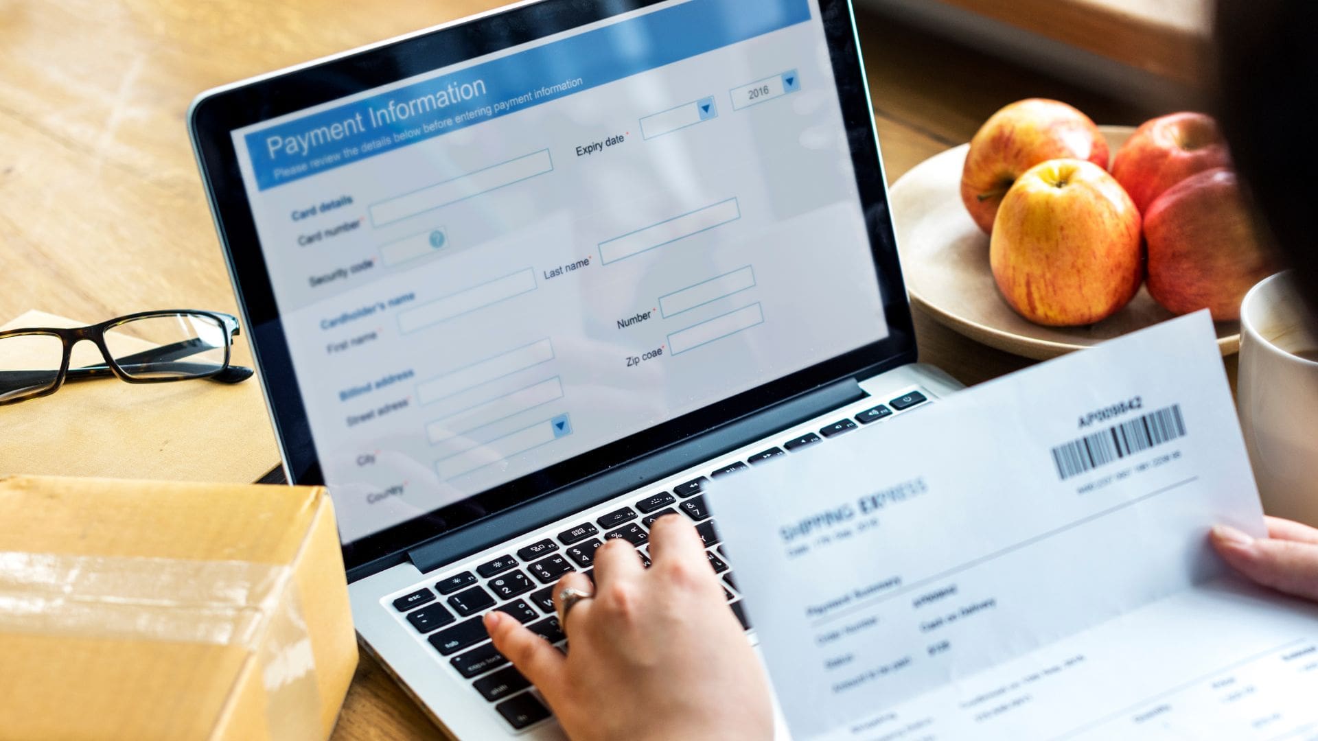 a hand holding invoice or billing statement to fill in computer laptop for shipping and billing details connection homepage with a spec on the muji style table and fruits on plate