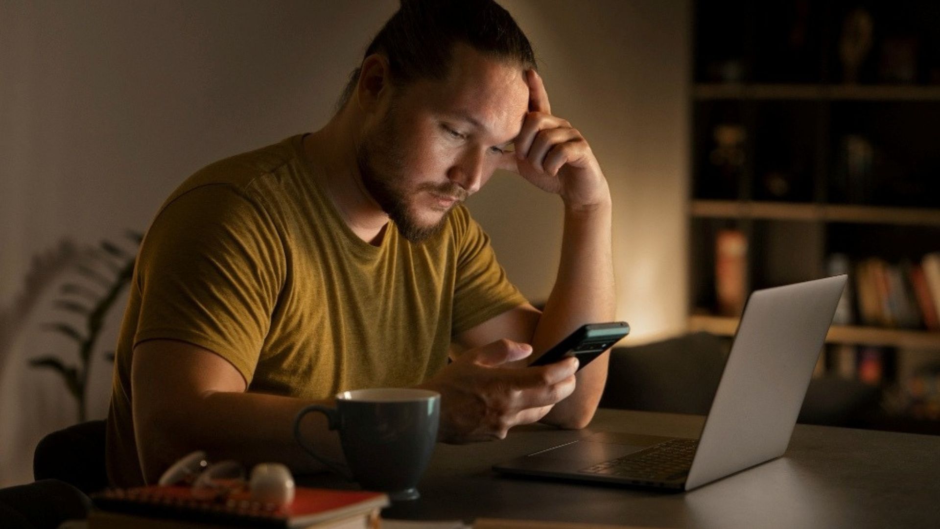 Image showing a man sitting at a desk late at night, looking concerned while checking his phone beside a laptop and coffee, representing an online business owner troubleshooting Shopify payment gateway issues.