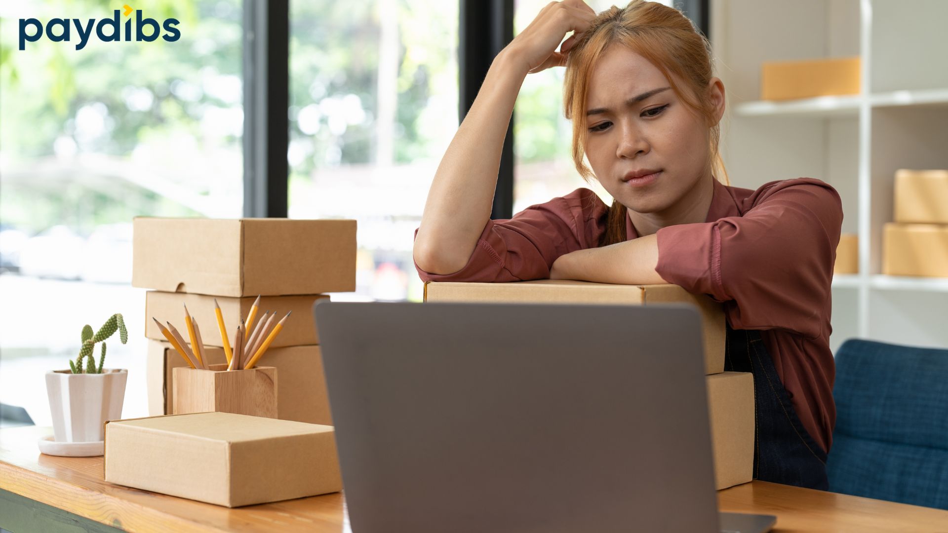 Paydibs branded image showing a small business owner sitting at a desk with parcel boxes and a laptop, looking stressed while managing online orders, illustrating common Shopify payment gateway problems faced by eCommerce sellers in Malaysia.